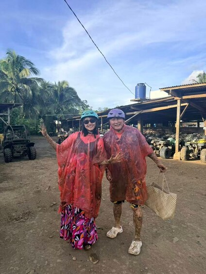 Two people covered in red paint, standing in a rural area with vehicles and palm trees in the background. 4