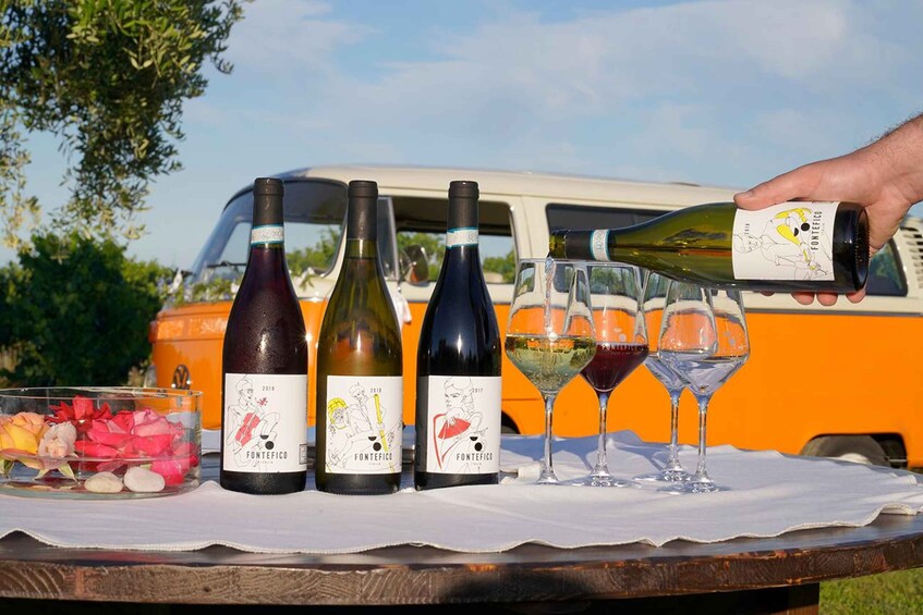 A wine tasting event with bottles and glasses on a table, a van in the background, and a person pouring wine. 1