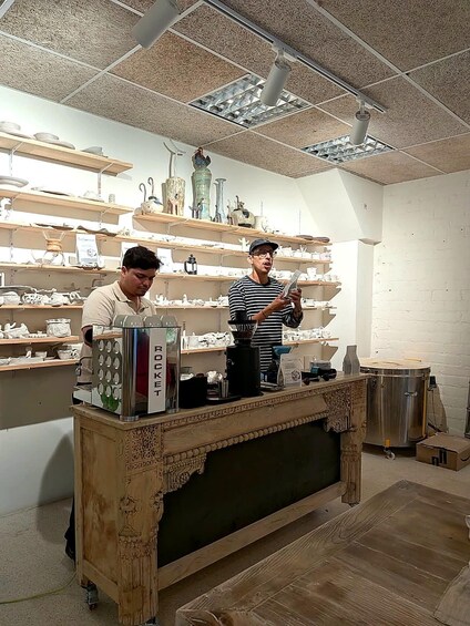 Two people in a pottery workshop with shelves of pottery and a coffee machine. 2
