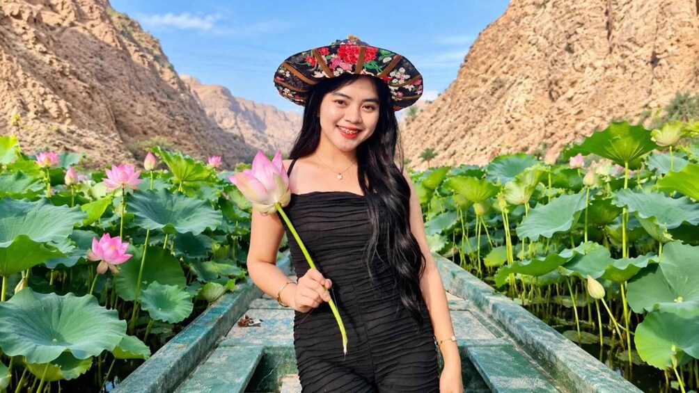 Una persona con vestido negro sosteniendo una flor de loto, de pie frente a un estanque de lotos con montañas al fondo. 1