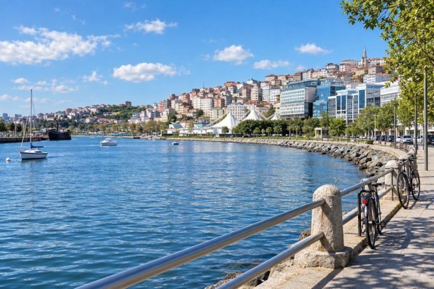 A waterfront with boats, a cityscape, and a paved walkway with a railing. 2