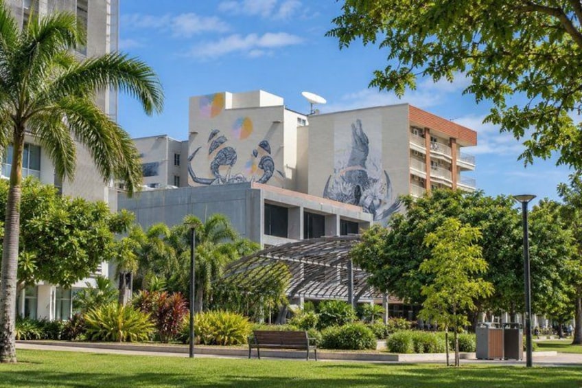 A building with murals, a park with trees and a bench, and a clear sky. 3