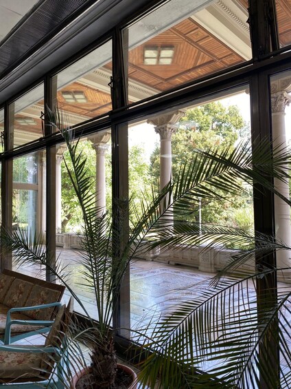 A conservatory with large glass windows, a wooden ceiling, and a view of a courtyard with palm trees and classical architecture. 2