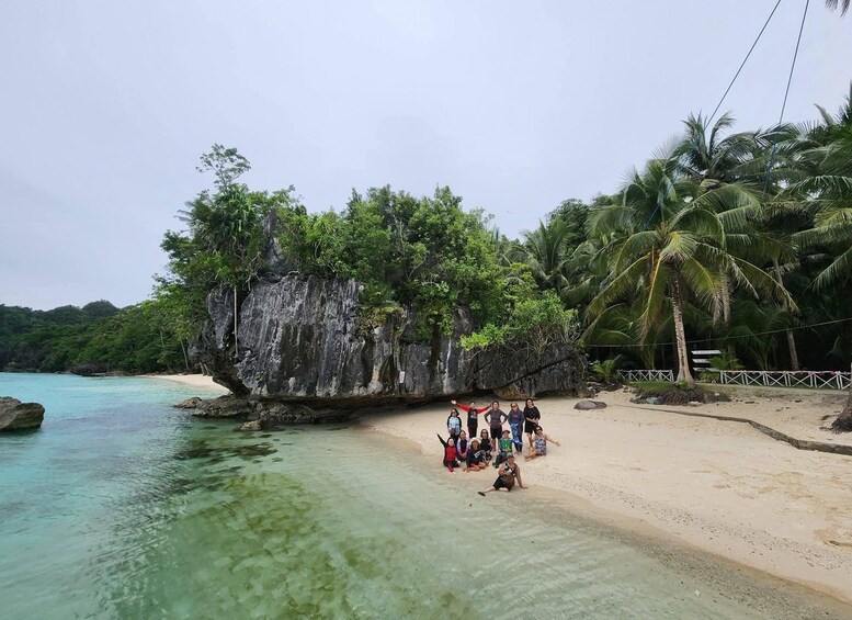 A beach with clear turquoise water, a large rock formation covered in greenery, and palm trees. 1