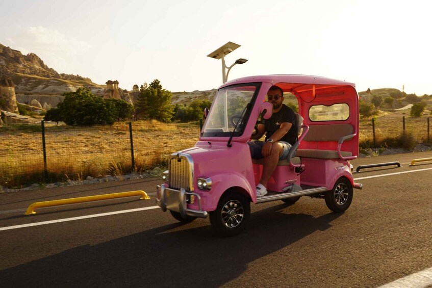A pink three-wheeled vehicle with a passenger, driving on a road with yellow barriers, in a hilly landscape with sparse vegetation. 3