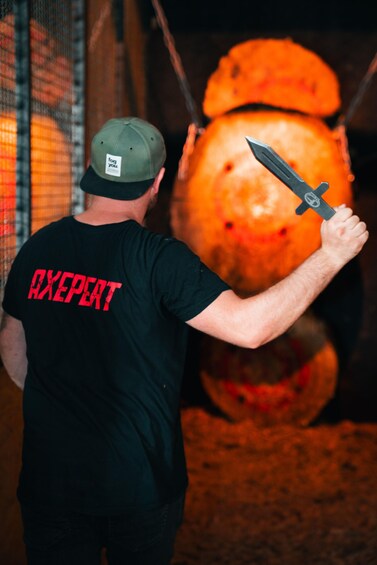 En person i en svart t-shirt med texten ”AXPERT” på ryggen, som håller en kniv framför ett stort orange föremål. 5