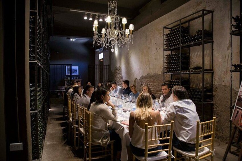A group of people seated around a round table in a dimly lit room with a chandelier, wine racks, and a brick wall. 4