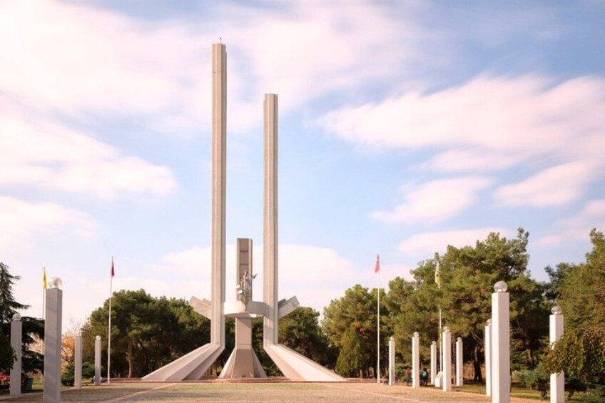 A memorial with tall, slender columns and a central sculpture, surrounded by trees and a clear sky. 3