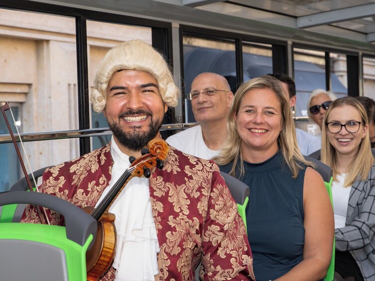 A person in a wig and patterned jacket holding a violin, standing in front of a vehicle with green seats. 1