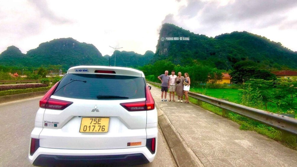 A white car with a yellow license plate on a road with a metal guardrail, surrounded by lush green hills and a cloudy sky. 5