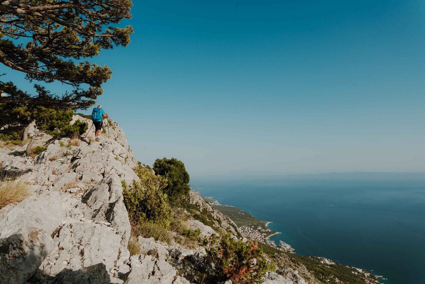 A hiker on a rocky mountain path with a view of the sea and a coastal town. 1