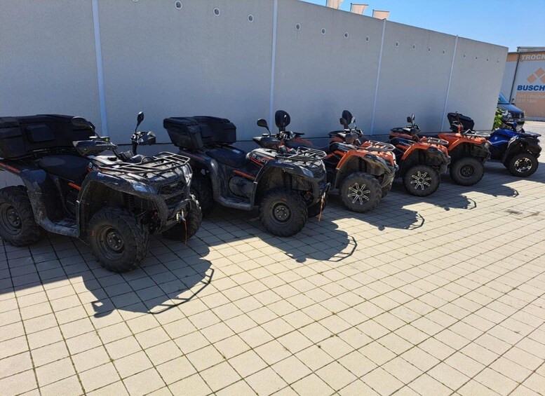 A row of ATVs parked in a lot with a white wall and a blue building in the background. 4