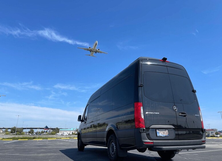 A black van parked in a lot with a clear blue sky and a plane flying overhead. 2