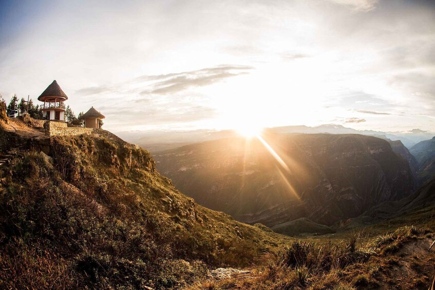 A mountain landscape with a small hut on a cliff, a valley below, and the sun shining through the clouds. 5
