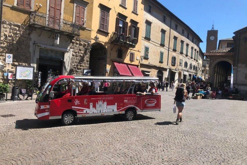 A red bus parked on a cobblestone street in a historic town square with buildings and a clock tower in the background. 4