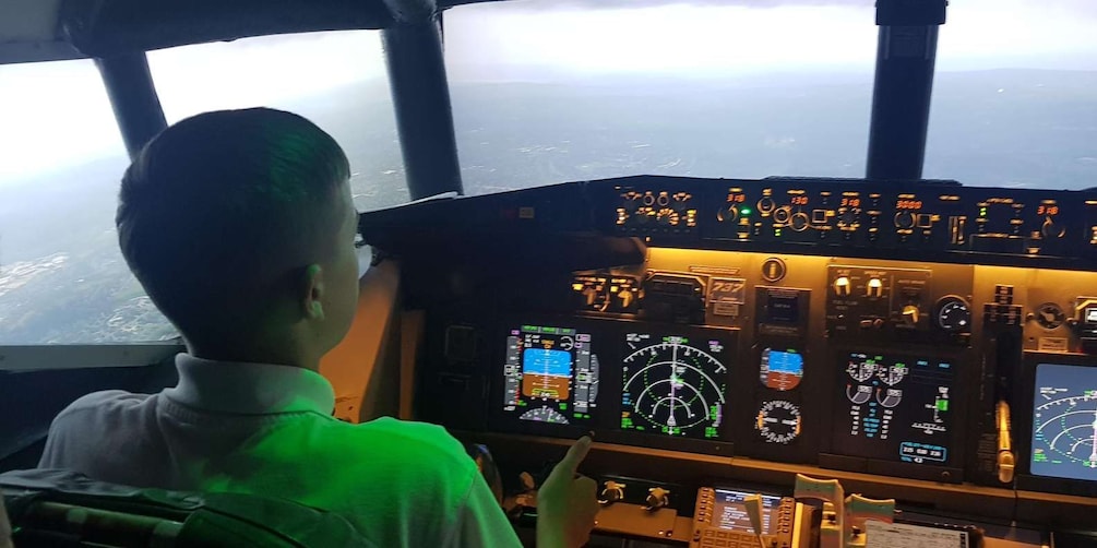 A cockpit with multiple screens and controls, a person pointing at the instruments, and a view of the sky through the windows. 1