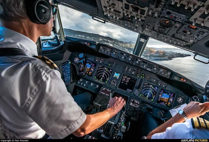 A pilot in a cockpit with multiple control panels and a view of a bridge and water outside. 4