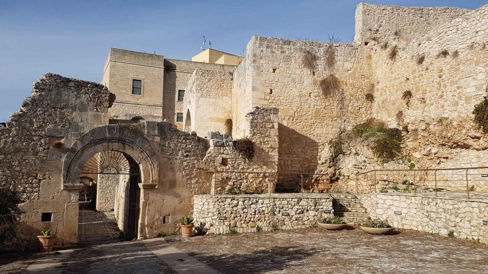 An ancient stone archway leading into a courtyard with a stone wall and a building in the background. 1