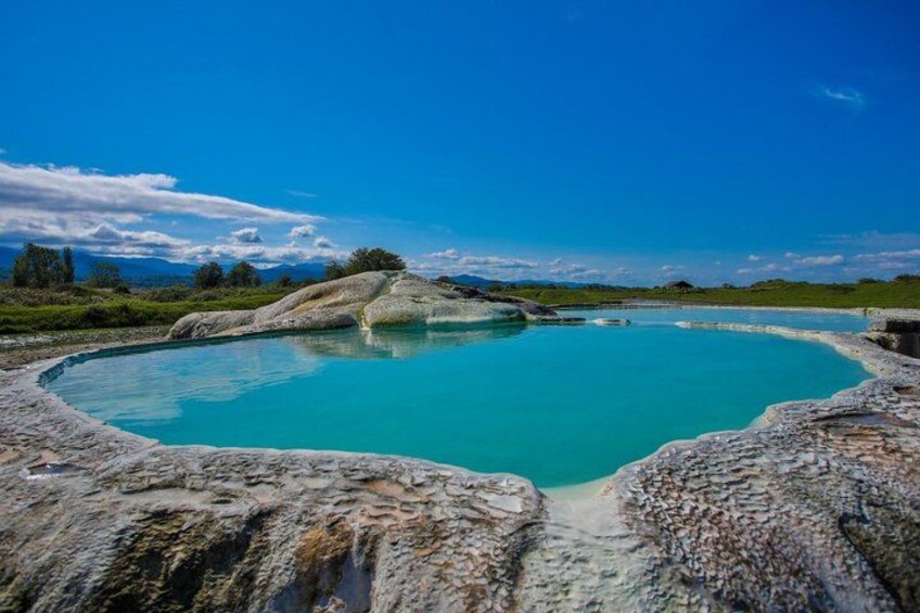 A turquoise hot spring surrounded by rocky terrain and a clear blue sky. 1