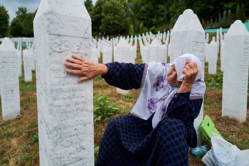 A person sitting on the ground, leaning against a white headstone in a cemetery. 1