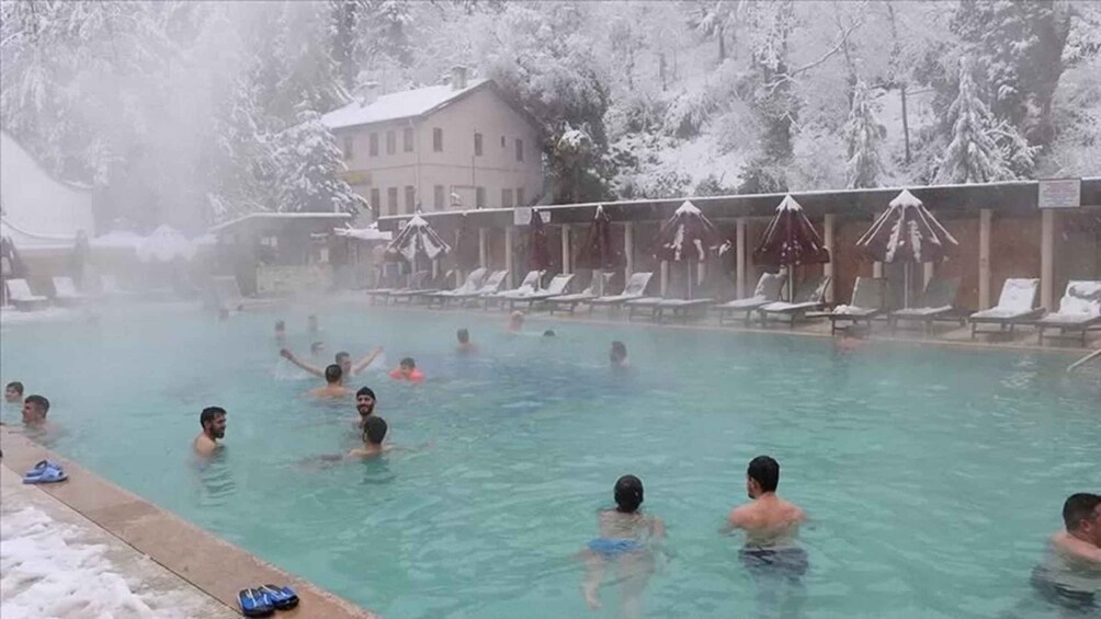 A swimming pool with people in the water, surrounded by snow-covered trees and a building. 1