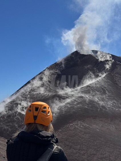 A person wearing a helmet and jacket observing a volcano with smoke or steam rising from its peak. 1