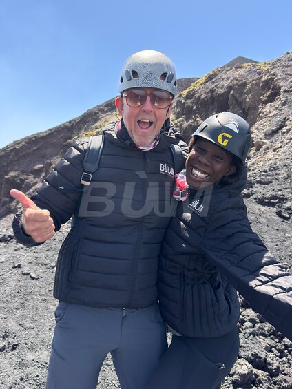 Two people wearing helmets and jackets, giving a thumbs up, standing on a rocky terrain with a clear sky. 3