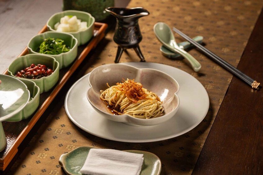 A bowl of noodles with toppings, accompanied by small bowls of condiments and a pair of chopsticks on a wooden table. 2