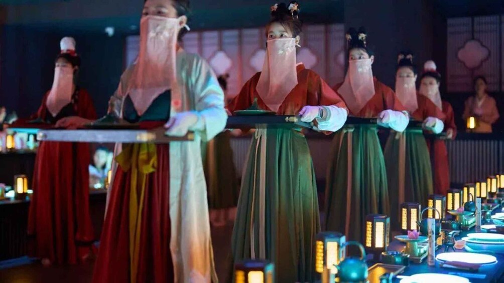 A group of people in traditional attire, holding a long object, possibly a ceremonial item, in a dimly lit room with tables set for a meal. 3