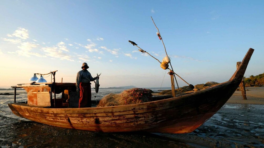 A wooden boat on a beach with a person standing on it, holding a fish. 3