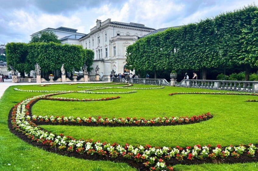 A formal garden with geometric flower beds, a large building, and a tree-lined pathway. 3
