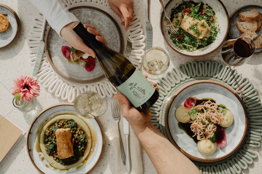 A person holding a bottle of wine, a plate with food, and a glass of wine on a table. 1