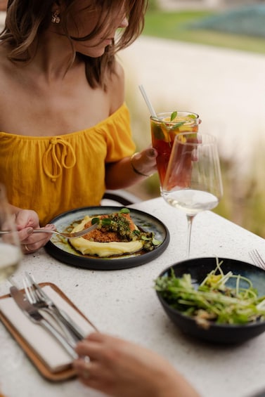 A person in a yellow off-shoulder top dining at an outdoor table with a meal, a drink, and a glass of white wine. 3