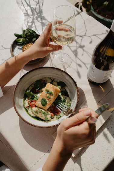 A person holding a glass of white wine, a fork, and a bowl of food with a piece of salmon. 5