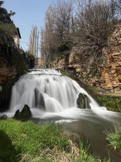 Ein Wasserfall stürzt über Felsen, umgeben von Grün und einem klaren Himmel. 1