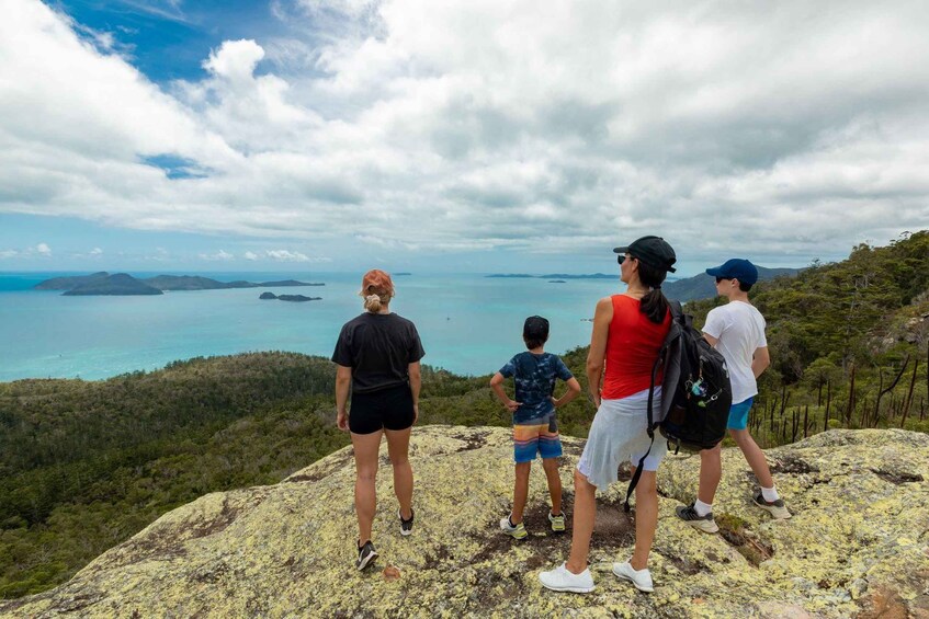 En familie på fire står på en klippe med udsigt over et kystlandskab med klare himmel og fjerne øer. 2