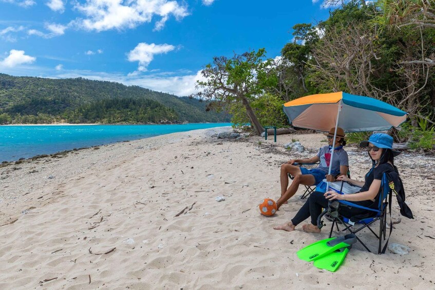 To personer sidder på strandstole under en parasol, med en fodbold og grønne flydeveste på en sandstrand med en klar blå himmel og turkis hav. 5