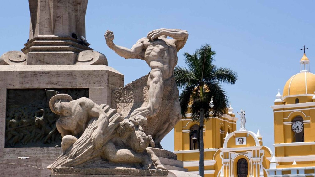 A large stone sculpture of a muscular figure with a raised arm, a palm tree, and a yellow building with a clock tower. 3