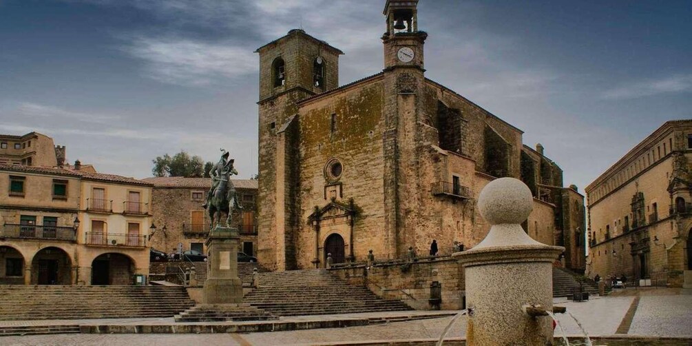 A historic church with a bell tower, a statue in front, and surrounding buildings. 1