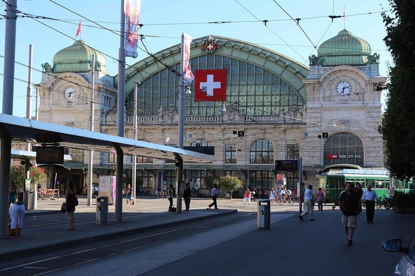 Ein Bahnhof mit einem hohen Glockenturm, einer Schweizer Flagge und einer Straßenbahn. 2