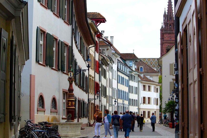 Eine enge Straße gesäumt von historischen Gebäuden, einem Brunnen und Spaziergängern. 4