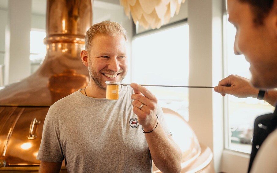 En person håller i ett litet glas med en bärnstensfärgad vätska, troligen en dryck, i ett rum där en stor koppardestillationsapparat syns i bakgrunden. 4