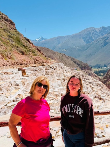 Two people standing in front of ancient stone ruins with mountains in the background. 1