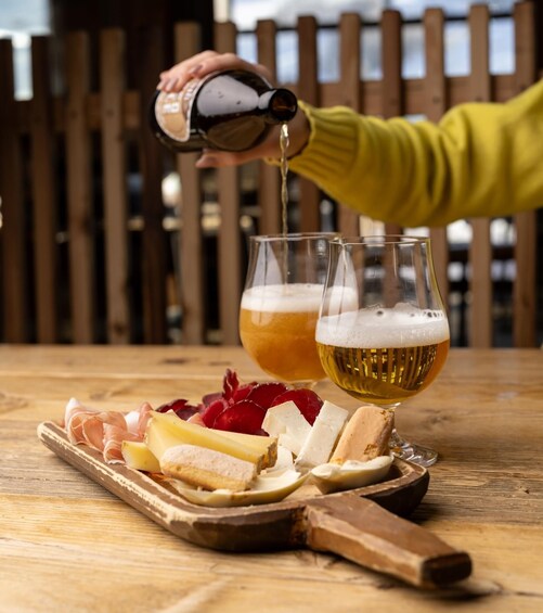 Une personne verse de la bière dans deux verres sur une table en bois, à côté de laquelle se trouvent du fromage et des fruits. 1