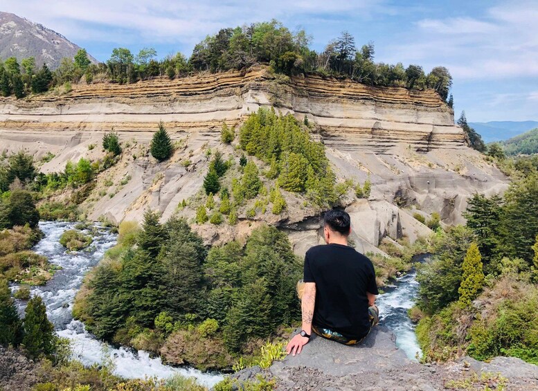 A man sitting on a rock overlooking a river with layered rock formations and dense greenery. 1