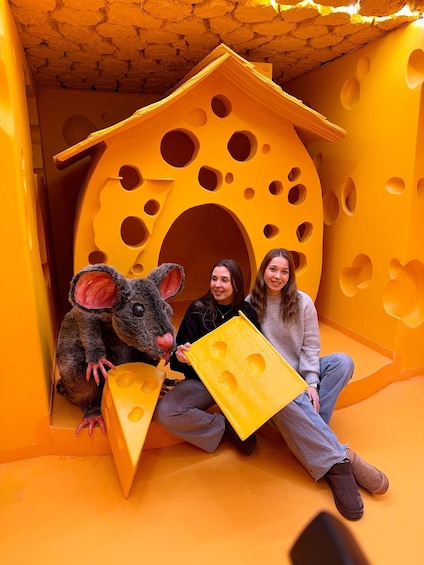 A large cheese sculpture with holes, resembling a mouse, and two people sitting in front of it. 3