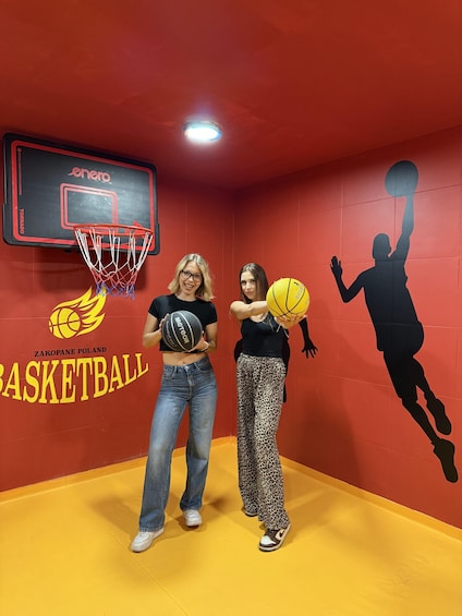 Two people holding basketballs in a gym with a basketball hoop and a wall with a basketball player silhouette. 2