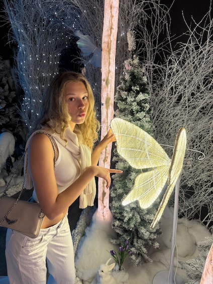 A person standing next to a decorative butterfly with lights, surrounded by snow-covered plants and a tree. 5