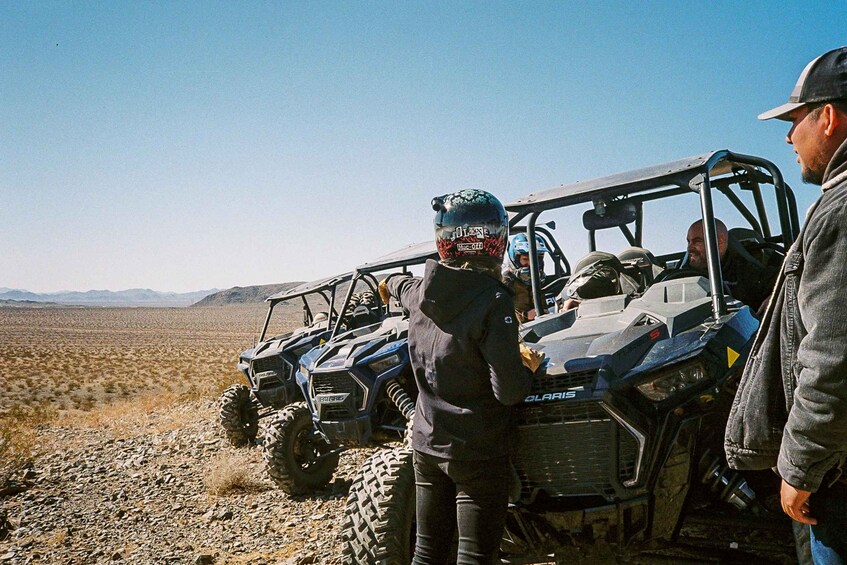 A group of people standing beside off-road vehicles in a desert landscape with mountains in the distance. 1