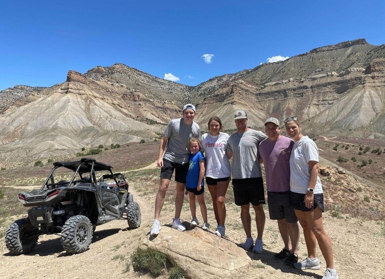 A group of people standing on a rocky outcrop with a quad bike in the foreground and a mountainous landscape in the background. 2